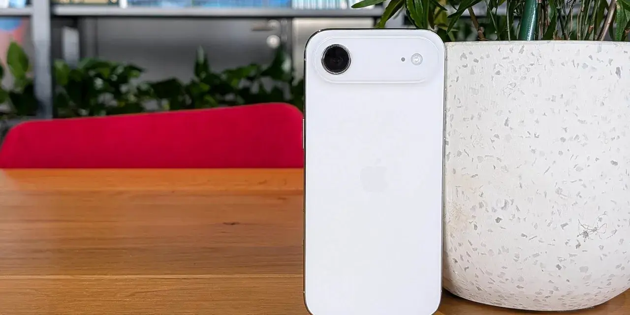 Biały iPhone z 4 aparatami, leżący na drewnianym blacie obok doniczki.