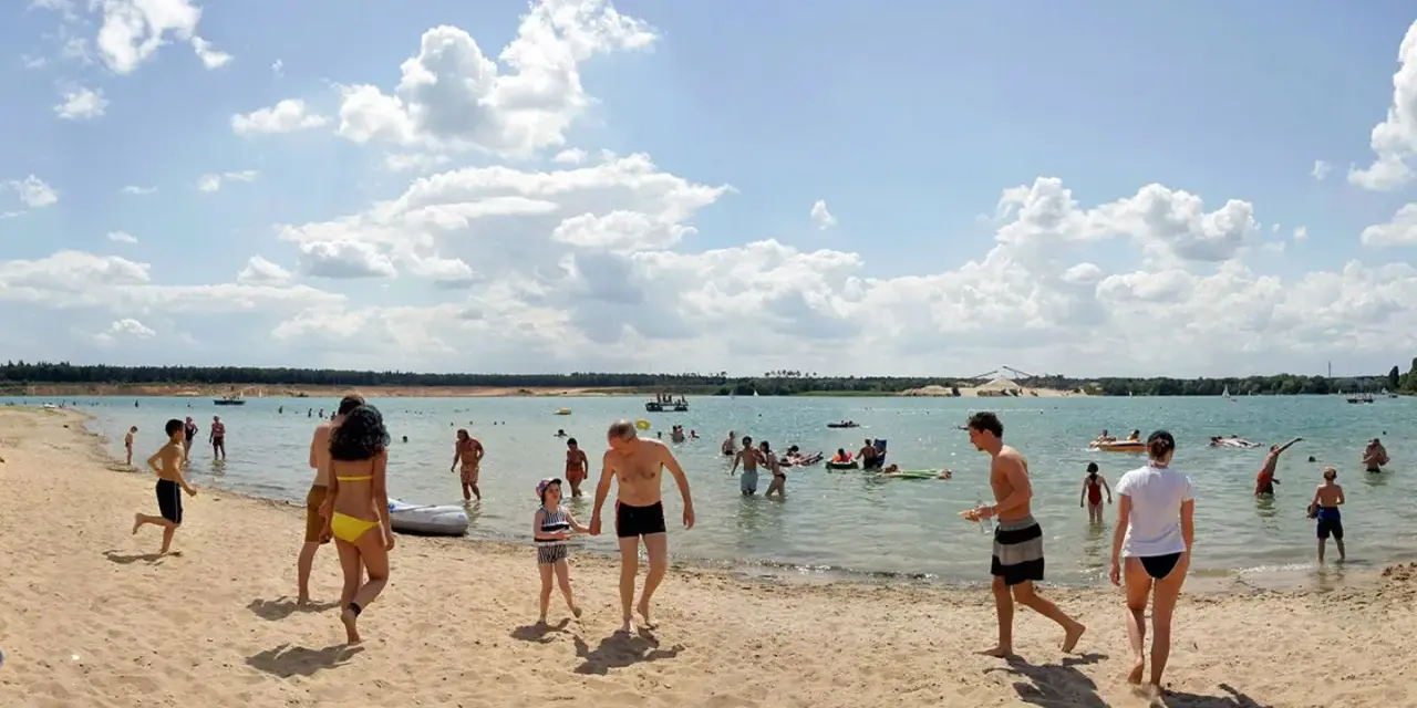 Menschen genießen einen sonnigen Tag am Badesee Frankfurt. Kinder planschen im Wasser, während Erwachsene sich am Sandstrand entspannen.