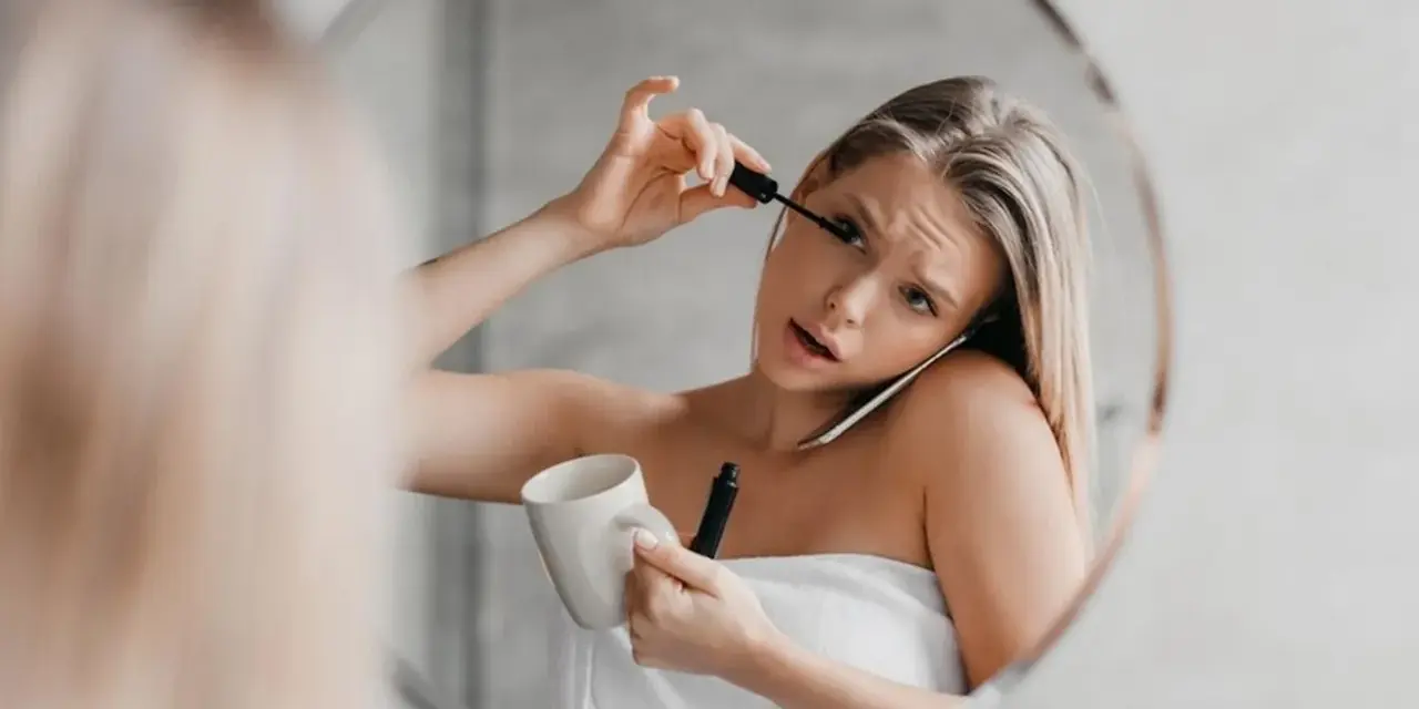 Une femme, enroulée dans une serviette, applique son mascara tout en tenant une tasse et en parlant au téléphone. Le mascara qui coule sur son visage témoigne de sa matinée mouvementée.