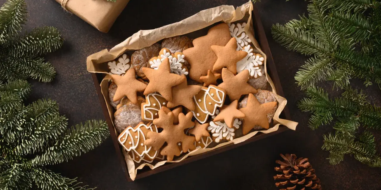 Last minute weihnachtsgeschenke selber machen: Eine Schachtel voller liebevoll verzierter Lebkuchen in Stern- und Baumform, umgeben von Tannenzweigen und Kiefernzapfen.