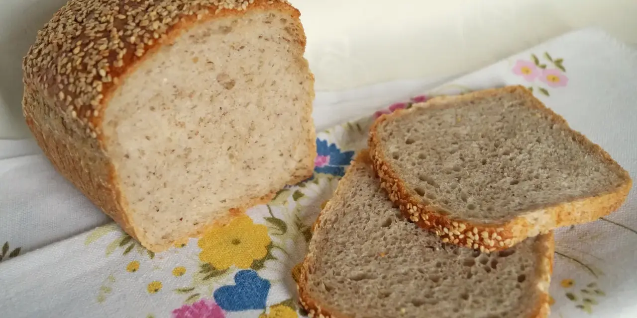 Chleb z kaszą gryczaną, posypany sezamem, leży obok dwóch kromek na kwiecistym obrusie.