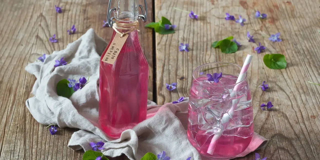 Flasche mit Veilchensirup und ein Glas mit Eis und Veilchenblüten auf Holzhintergrund.