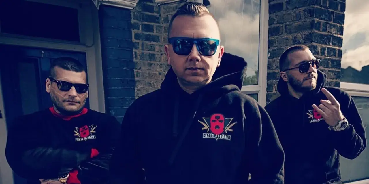 Trzech mężczyzn w bluzach z logo "Gang Albanii". Jeden z nich pokazuje gest ręką.