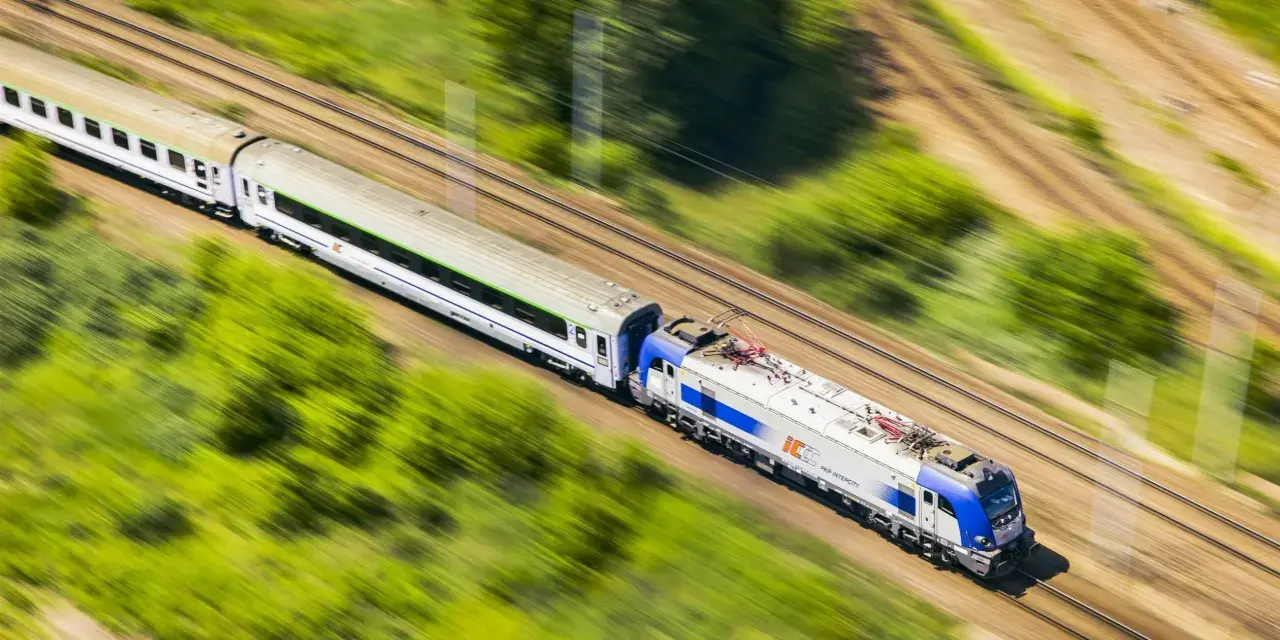 Pędzący pociąg PKP Intercity, model EU44 "Husarz", z białymi wagonami, mija zielone pola i drzewa.