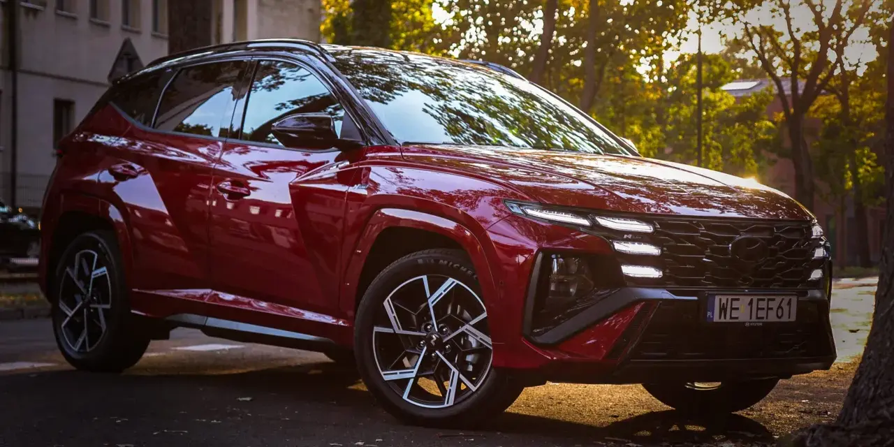 Czerwony Hyundai Tucson Hybrid zaparkowany na tle jesiennych drzew i kamienicy.