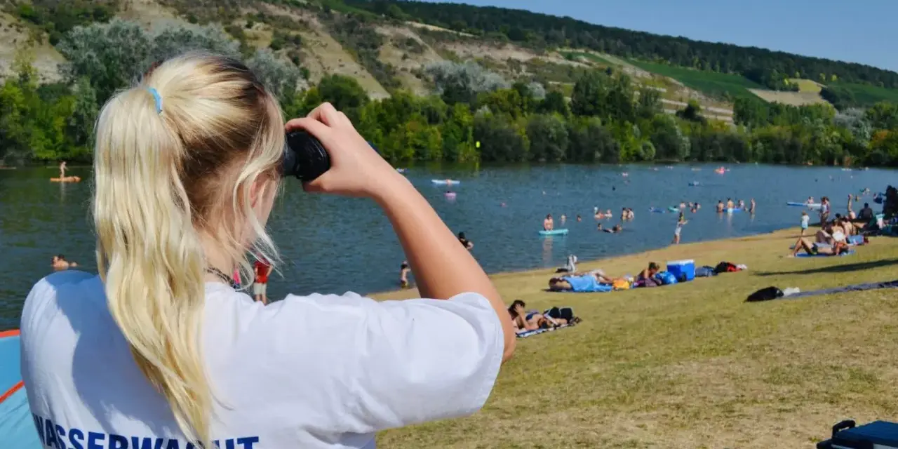 Wasserwacht am Erlabrunner Badesee: Eine junge Frau mit blondem Pferdeschwanz blickt durch ein Fernglas über den belebten See, auf dem viele Menschen schwimmen und sonnen.