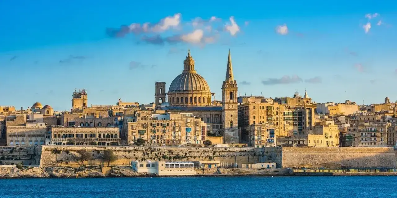 Malta, wyspa do kogo należy? Widok na Vallettę z kopułą i wieżą kościelną, otoczoną zabytkową architekturą, nad błękitnym morzem.