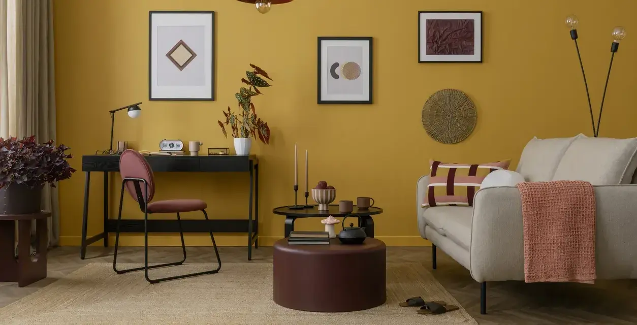 Un salotto con pareti giallo ocra e dettagli tortora. Un divano beige, un pouf tortora e un tappeto in juta creano un'atmosfera accogliente.