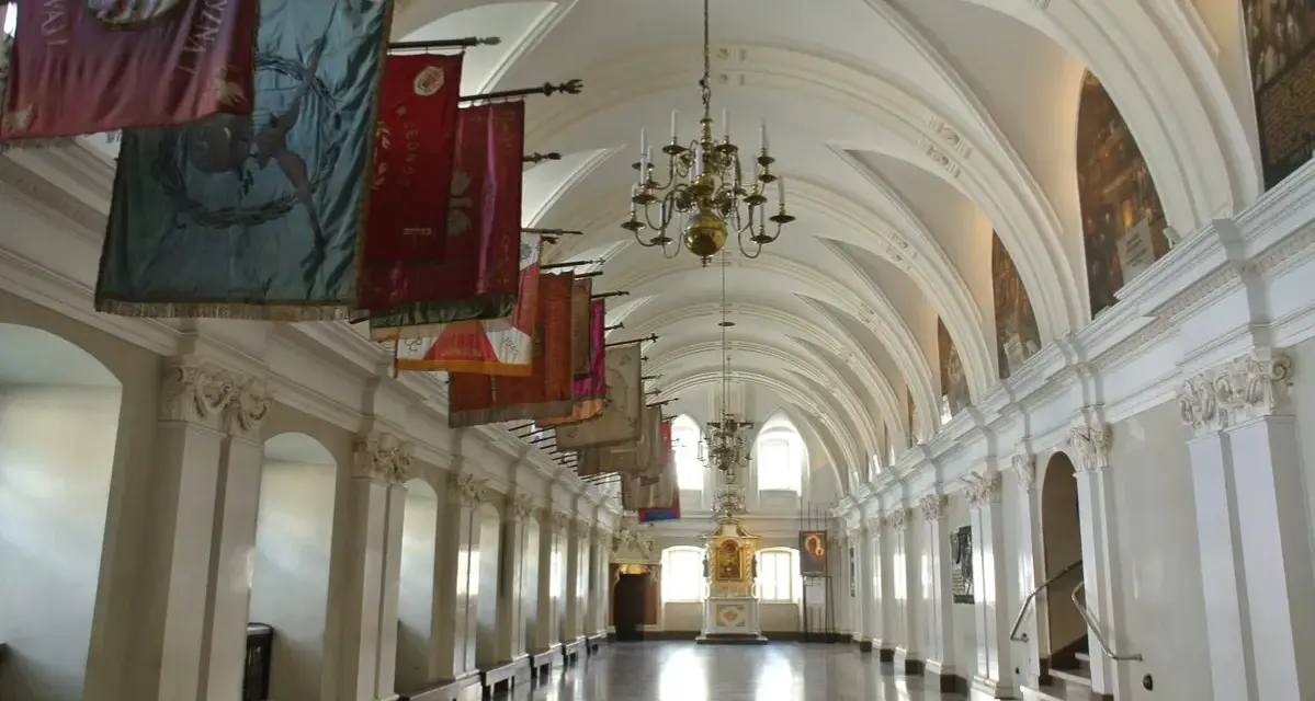 Długi korytarz z łukami i żyrandolami, ozdobiony historycznymi sztandarami. To wnętrze klasztoru na Jasnej Górze, gdzie każdy baner opowiada historię zakonu paulinów.