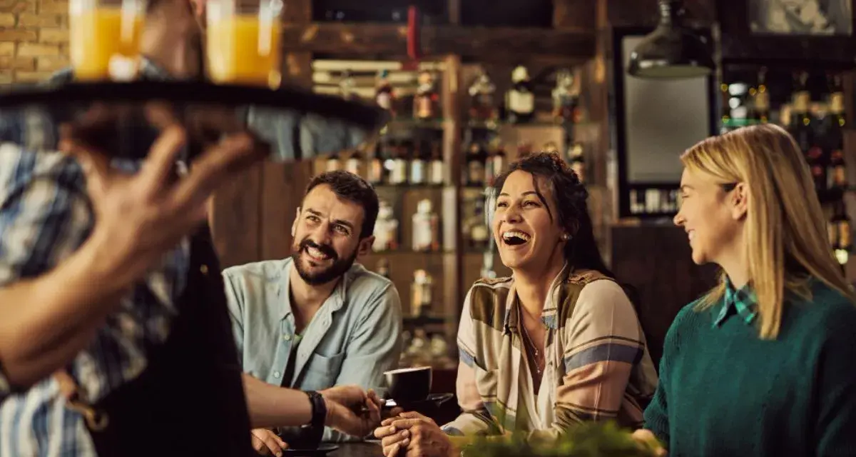 Un camarero sirve bebidas a un grupo de amigos riendo en un bar. La conversación animada sugiere que aún no es tarde para el ruido.