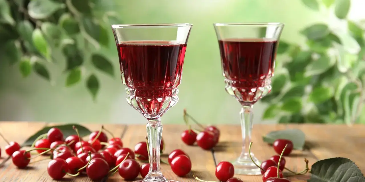 Dwa kieliszki z rubinową nalewką z wiśni na spirytusie, obok świeże wiśnie na drewnianym stole. Szybka nalewka z wiśni na spirytusie gotowa do degustacji.