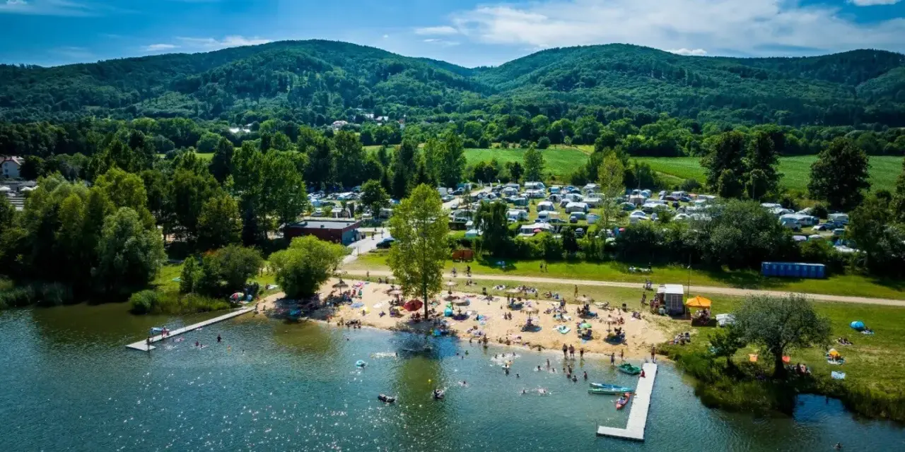 Słoneczny dzień nad jeziorem Bielawa. Tłumy plażowiczów, kemping z kamperami i zielone wzgórza w tle.
