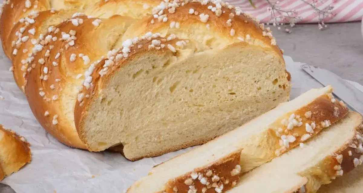 Ein goldbrauner Hefezopf, bestreut mit Zuckerkristallen, liegt auf Backpapier. Ein Stück ist angeschnitten, die weiche Krume ist sichtbar. Ein tolles Rezept für Hefezopf!