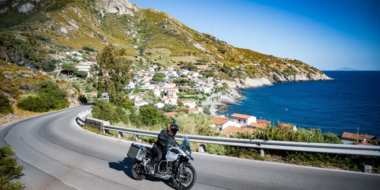 Un motociclista esplora le curve panoramiche dell'Elba, con il mare blu e un villaggio pittoresco sullo sfondo.