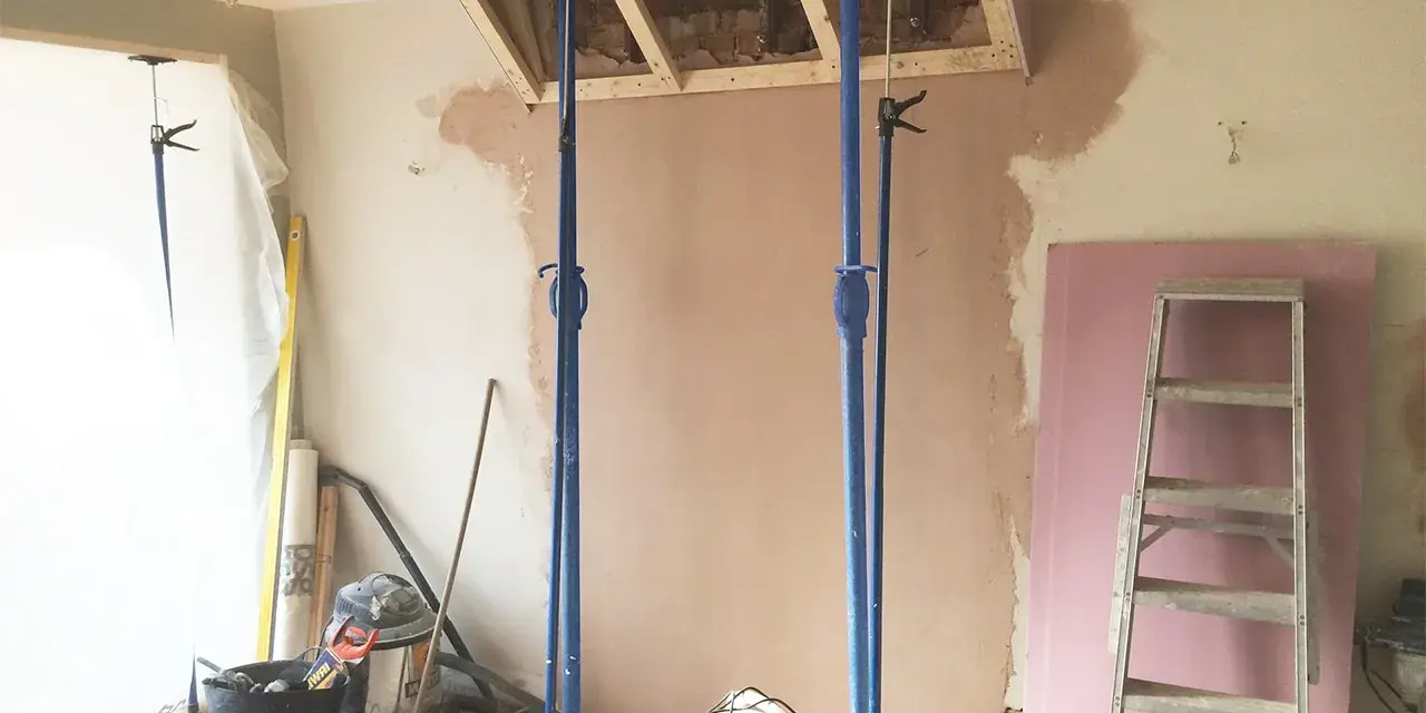 Travaux de rénovation : des étais soutiennent une structure en bois au-dessus d'une zone où l'on s'apprête à casser une cheminée. Matériaux de construction et outils au sol.