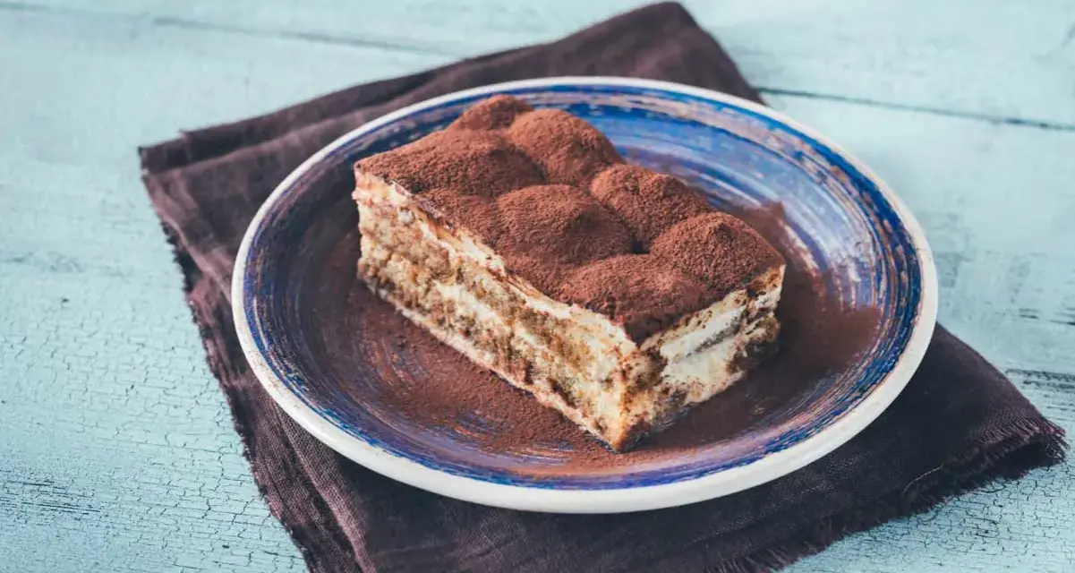 Pyszny kawałek prawdziwego tiramisu, posypany kakao, na niebieskim talerzu.