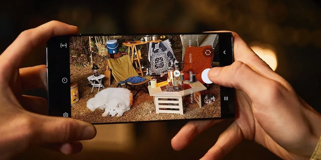 Une photo de téléphone capture un chien blanc endormi lors d'un camping. L'écran du téléphone montre une scène de camping avec une tente, une chaise pliante et un feu de camp.