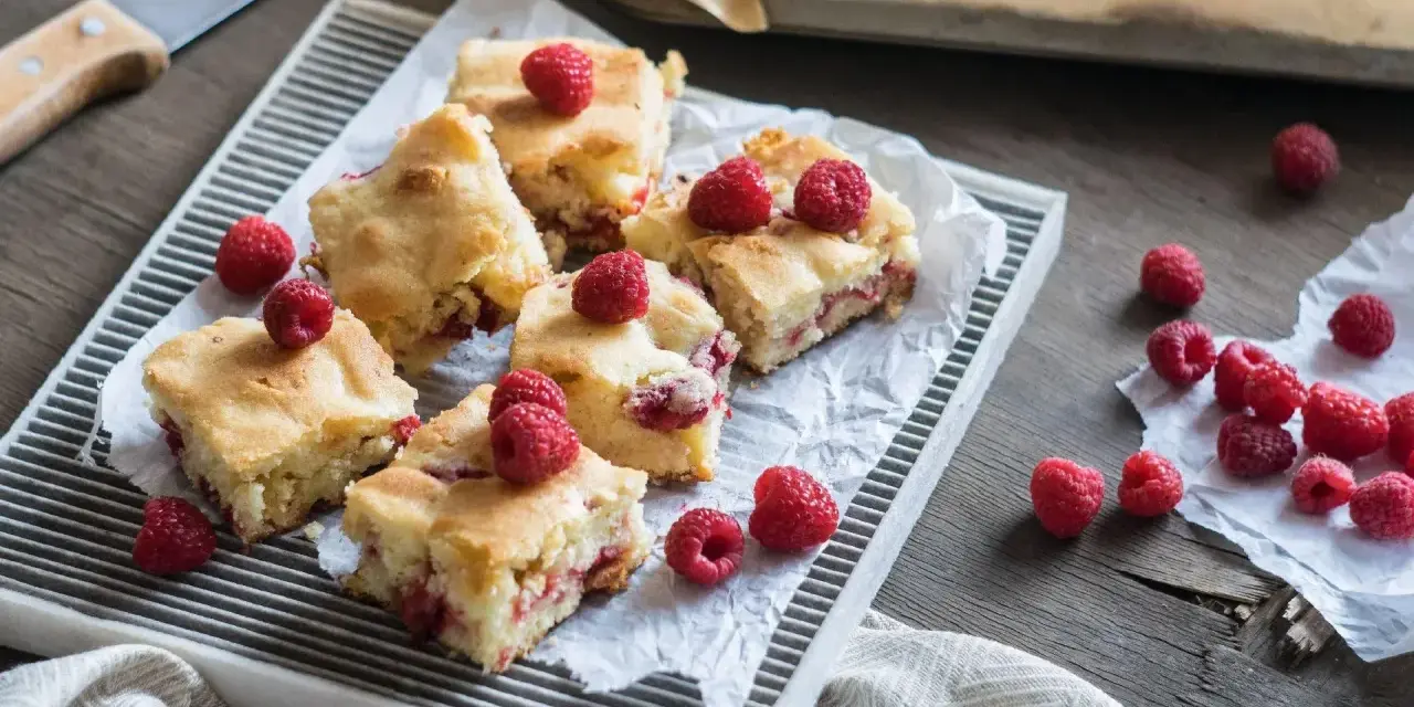 Słodkie brownie i blondie z malinami, pokrojone na kwadraty, ułożone na tacy. Kilka malin rozsypanych obok.