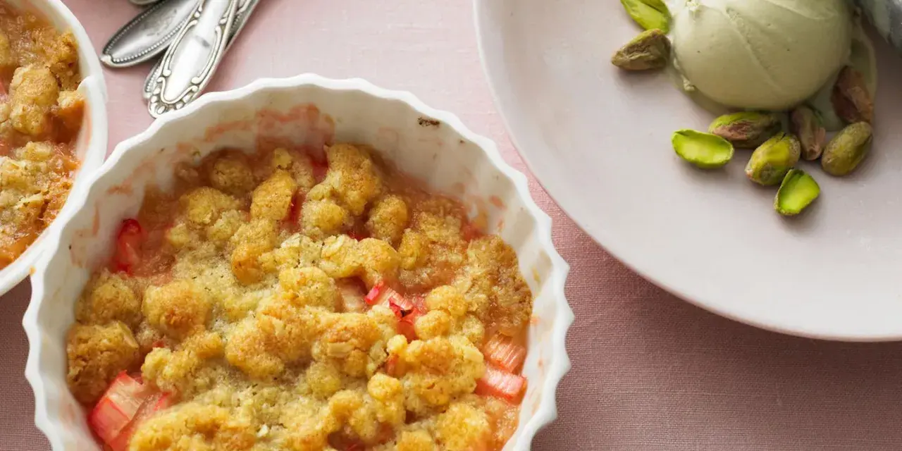 Rhabarber-Crumble in weißer Auflaufform, daneben eine Kugel Eis mit Pistazien. Ein köstliches Rhabarber Dessert.
