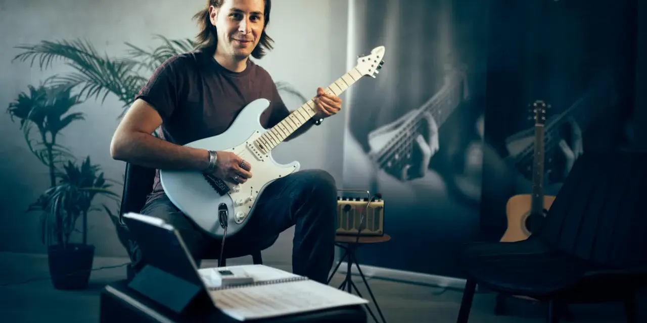 Mann mit E-Gitarre, bereit, online Gitarre zu lernen. Noten und Tablet liegen bereit.