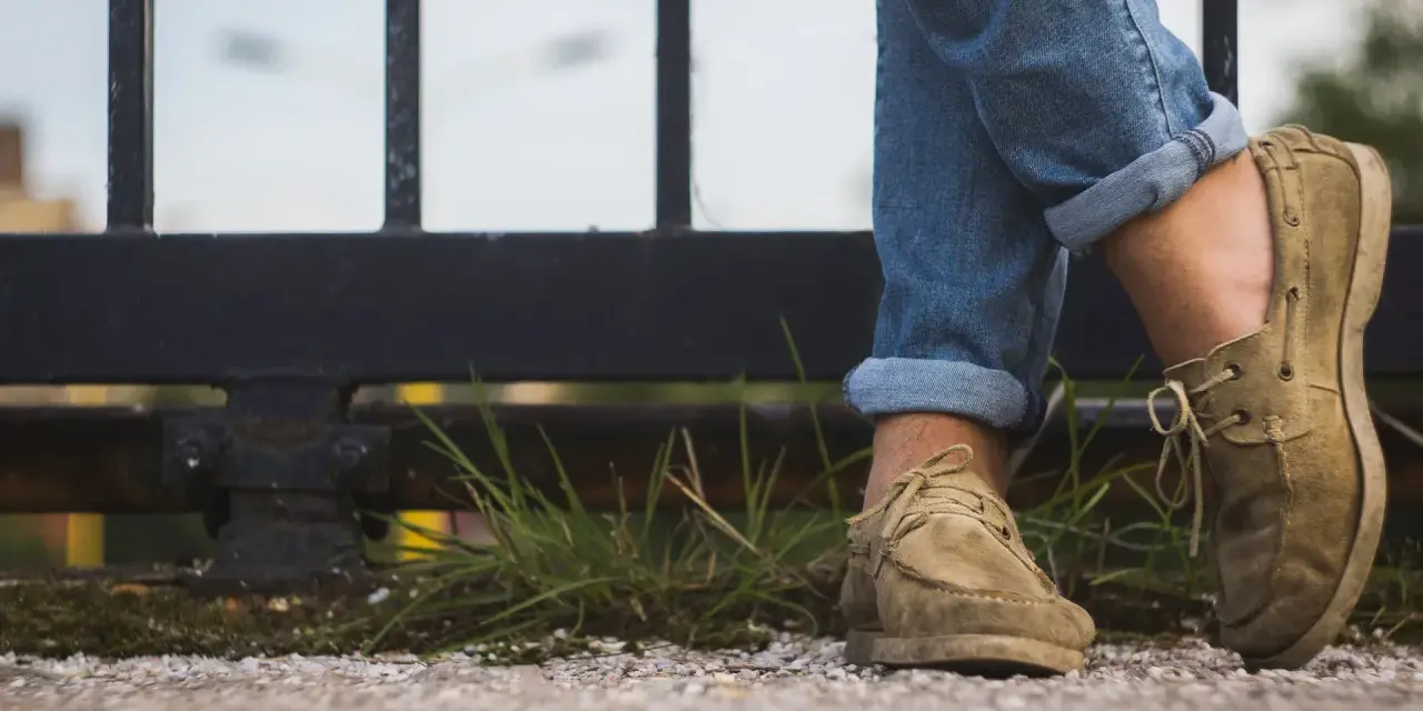 Gambe con jeans arrotolati e scarpe senza calzini, appoggiate su un muretto con erba.