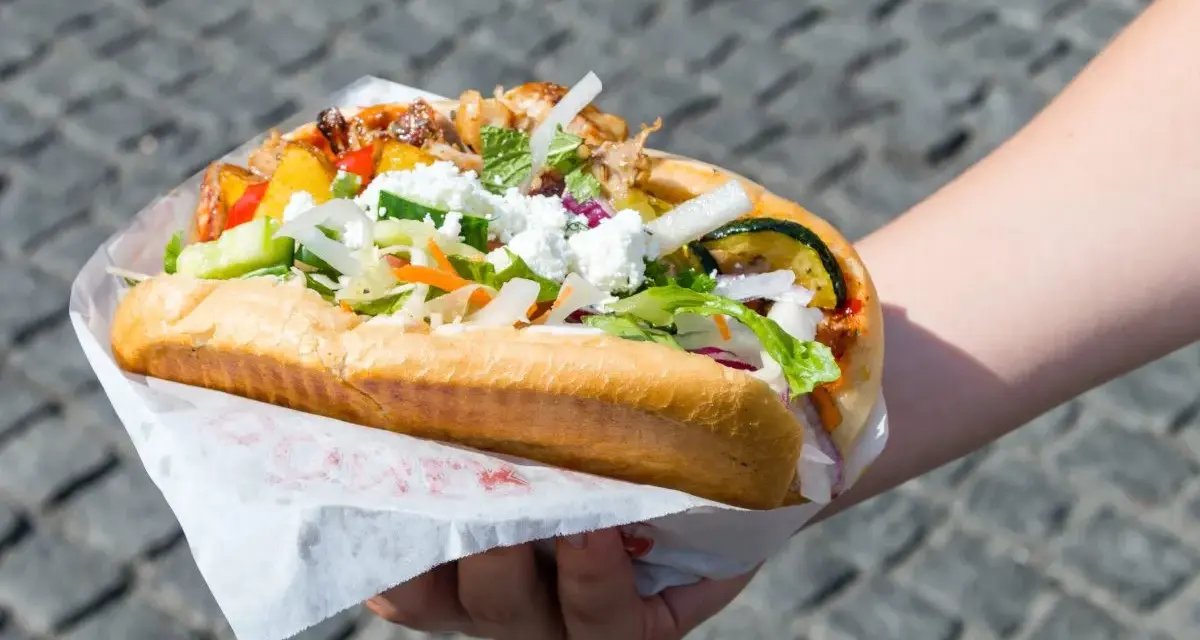 Kebab w bułce z mięsem, warzywami i serem feta, trzymany w dłoni.