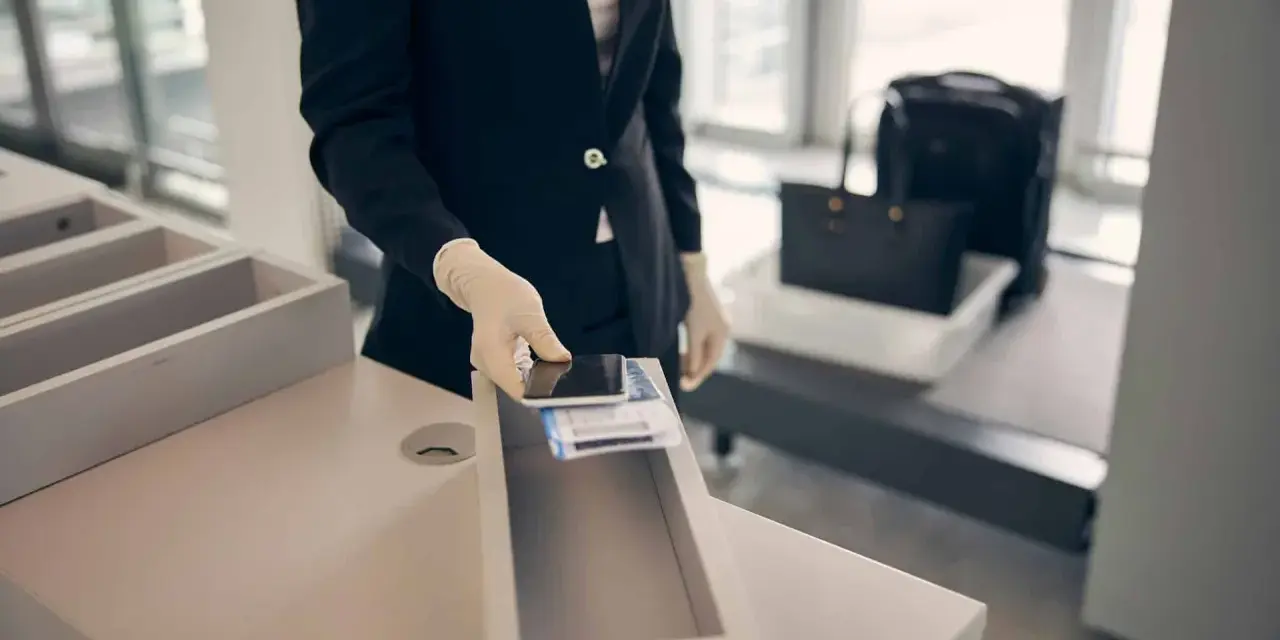 Pracownik lotniska w rękawiczkach kładzie telefon i karty pokładowe do pojemnika. W tle widać bagaż na taśmie. Zapalniczka w samolocie jest zabroniona.