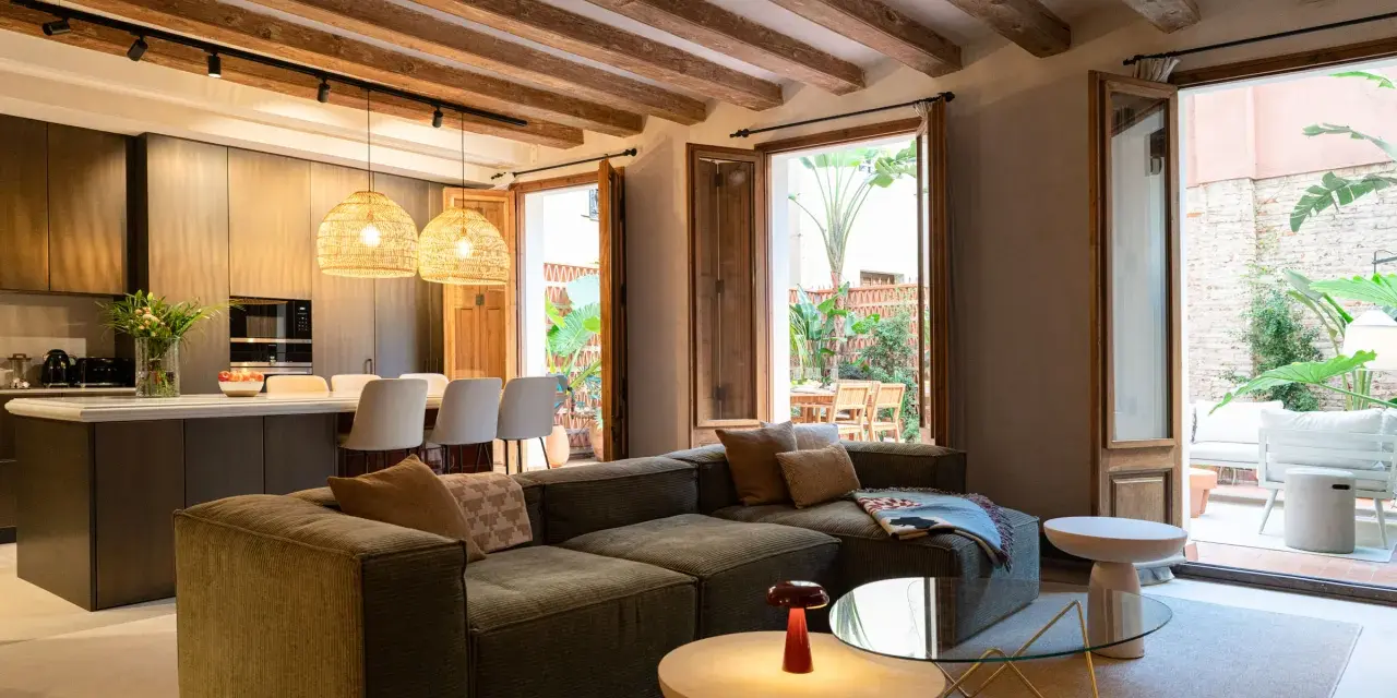 Sofá modular verde con acabado fratasado, mesas auxiliares, lámpara roja y acceso a patio con muebles de exterior.