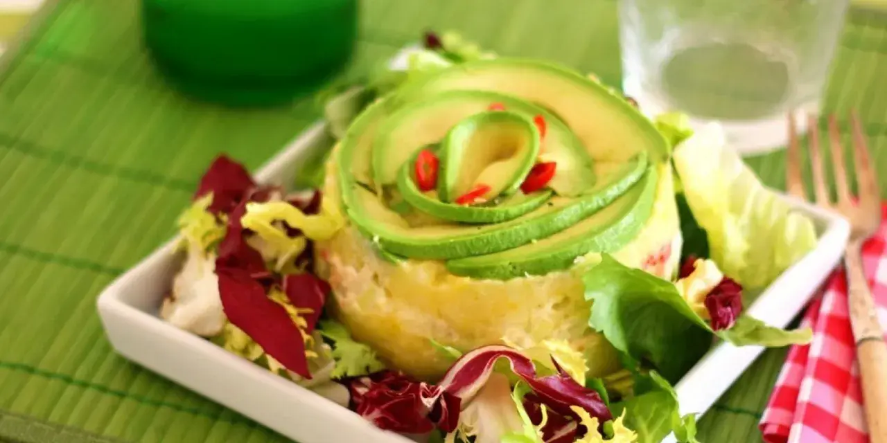 Causa limeña con aguacate en espiral y ensalada fresca, servida en plato blanco.