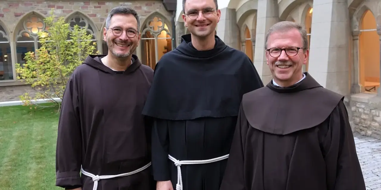 Drei Franziskaner Minoriten in braunen und schwarzen Kutten lächeln in die Kamera vor einer Klosterfassade.