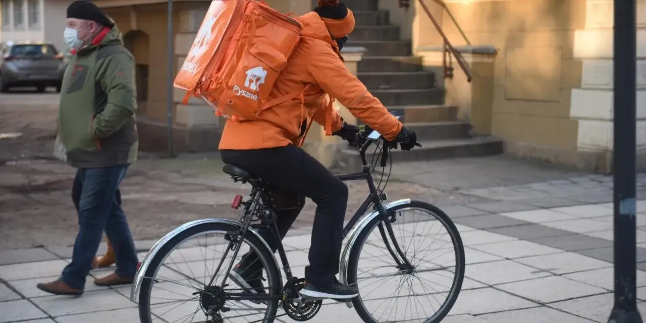 Kurier rowerowy w pomarańczowej kurtce z plecakiem Pyszne.pl, zarobki zależą od liczby dostaw.