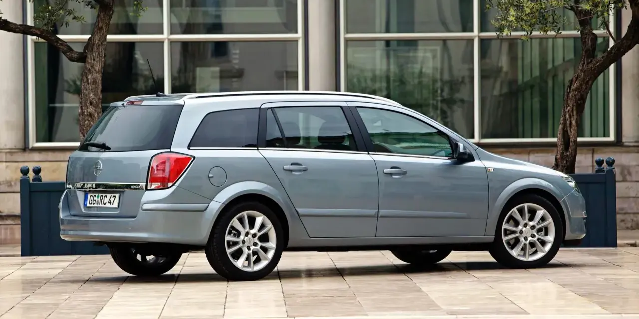 Srebrny Opel Astra H kombi zaparkowany przed nowoczesnym budynkiem z drzewami.