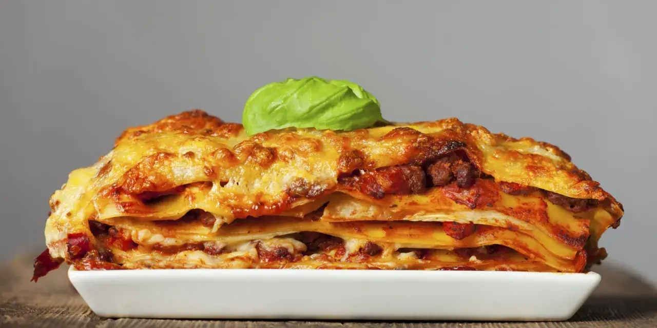 Una porción generosa de lasaña de carne, con capas de pasta, salsa y queso dorado, coronada con una hoja de albahaca fresca.