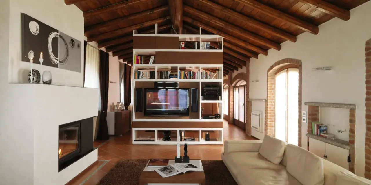 Accogliente soggiorno mansardato con soffitto in legno a vista, camino moderno e libreria centrale. Idee mansarde in legno per un ambiente caldo.