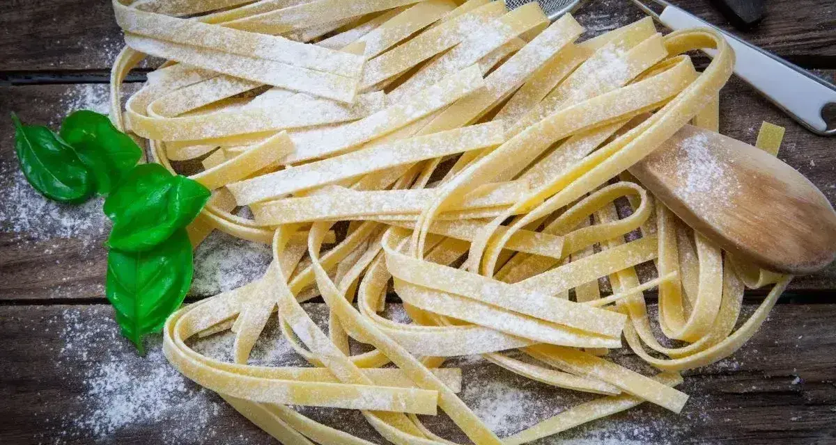 Świeży, domowy makaron bez glutenu, posypany mąką, z listkami bazylii i drewnianą łyżką.