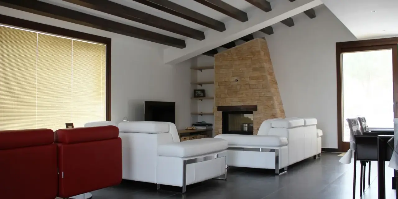 Soffitto con travi in legno scuro, un camino in pietra e divani bianchi. Ecco come fissare travi in legno al soffitto per un effetto rustico.