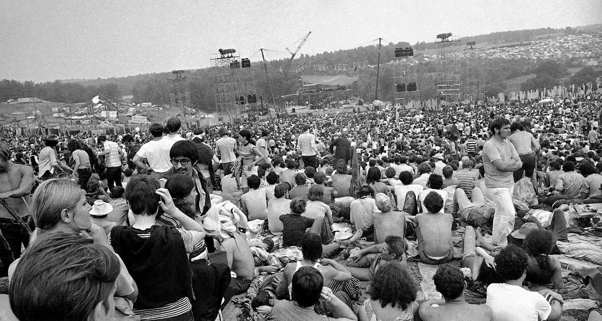 Tysiące ludzi siedzi na trawie, patrząc na scenę festiwalu. To jest Woodstock, gdzie jest muzyka i wolność.
