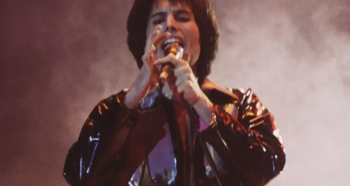 Freddie Mercury śpiewa z pasją, czując, że chce się uwolnić. Błyszcząca kurtka odbija światła sceny.