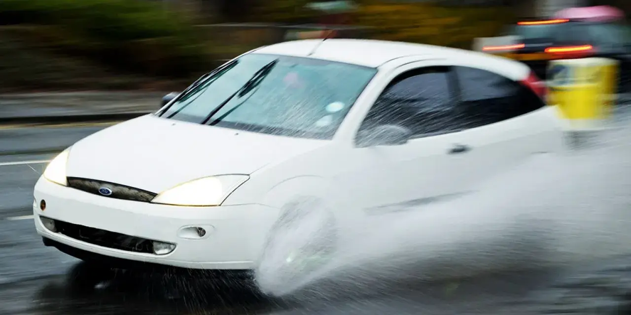 Auto bianco in movimento su strada bagnata, con un'enorme spruzzata d'acqua che indica l'aquaplaning cos'è.