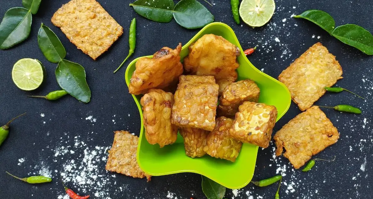 Knusprig gebratener Tempeh, ein fermentiertes Sojaprodukt, liegt in einer grünen Schale. Dazu garnieren Limettenscheiben, grüne Chilis und Blätter.