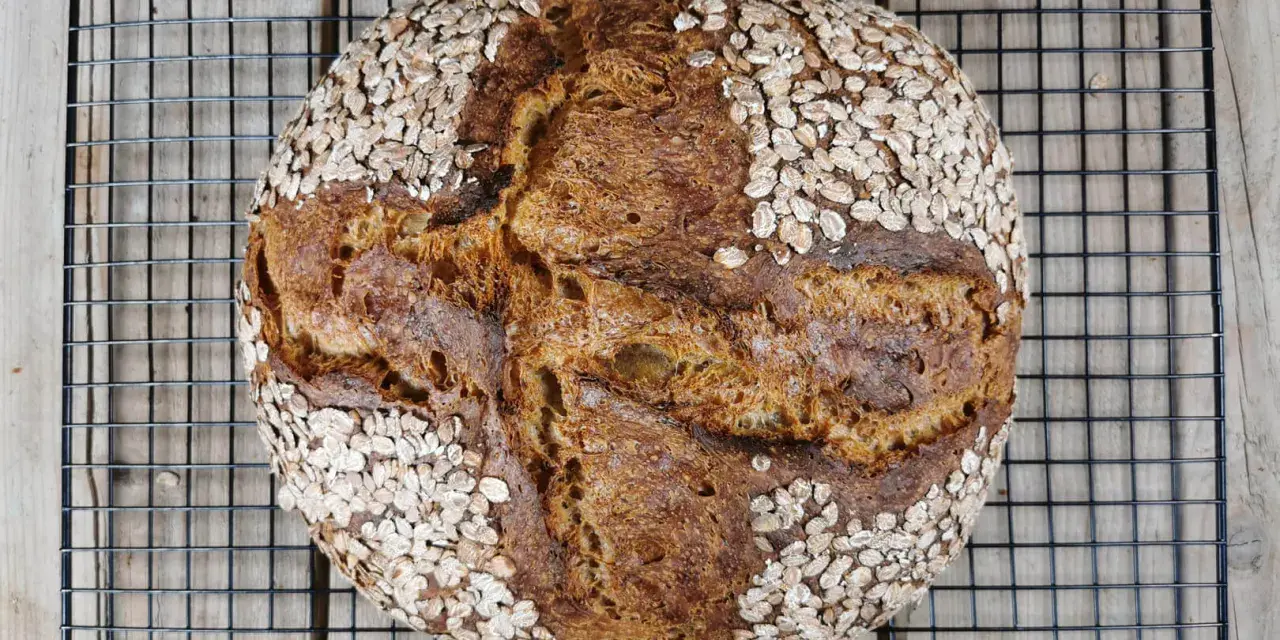 Ein rustikales Porridge Brot mit Haferflockenkruste, das auf einem schwarzen Gitter auf Holzdielen abkühlt.