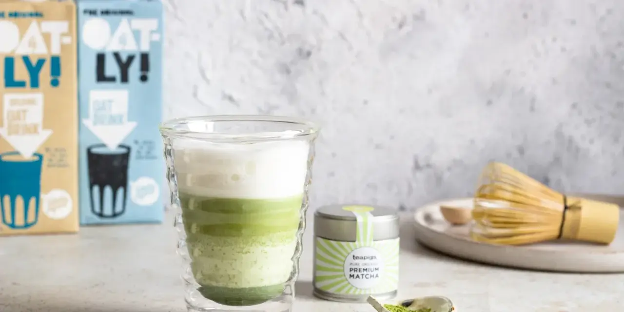 Warstwowy napój matcha z mlekiem kokosowym w szklance z podwójnymi ściankami. Obok puszka z matchą i bambusowa miotełka.