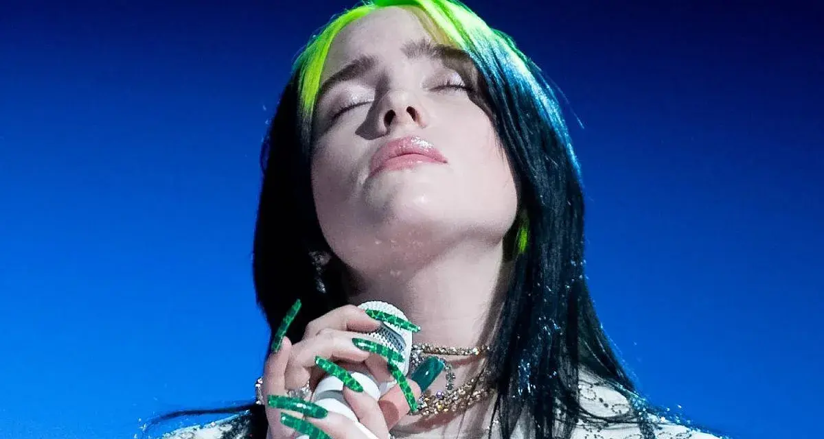 Piosenkarka Billie Eilish z zamkniętymi oczami śpiewa do mikrofonu, jej zielone pasemka włosów lśnią.