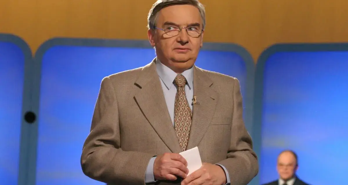 Tadeusz Sznuk, który prowadzi teleturniej Jeden z dziesięciu, w beżowej marynarce i z kartką w dłoni.