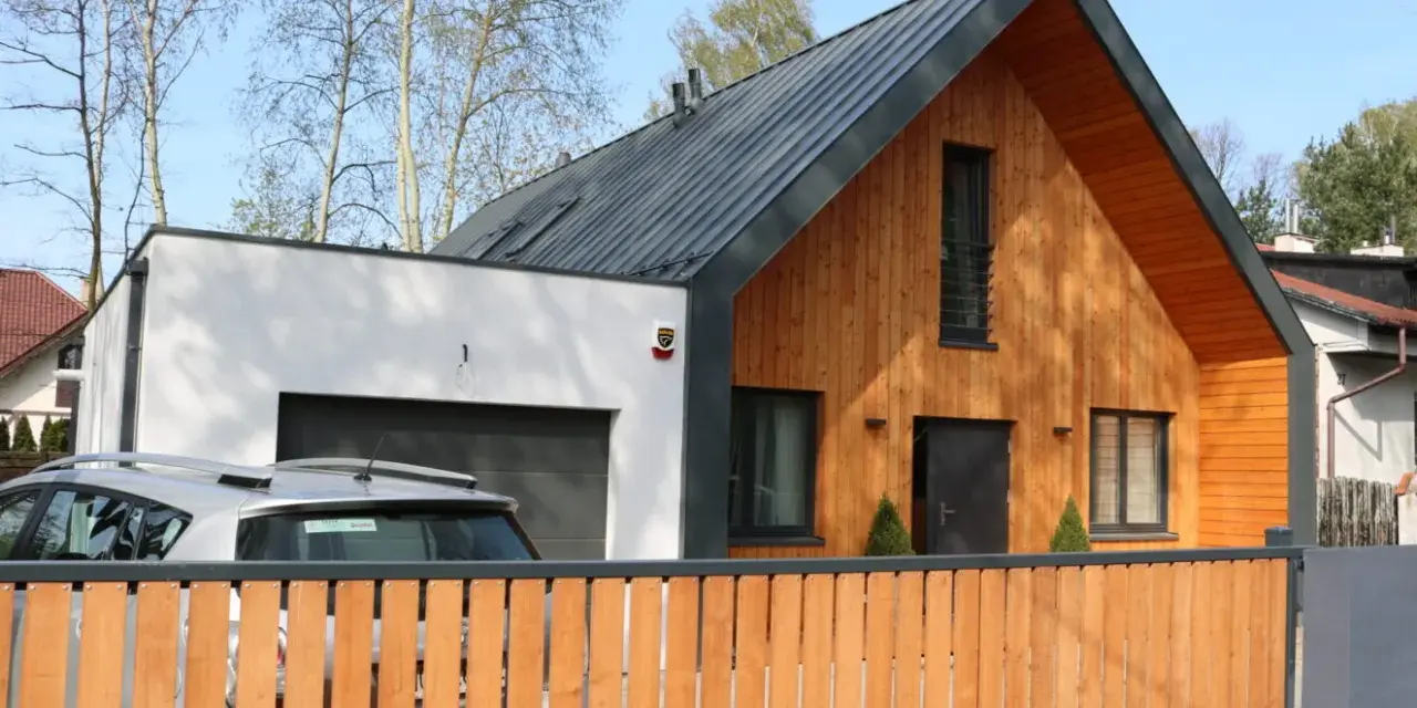 Nowoczesny dom z drewnianą dobudówką do części murowanej. Obok garaż, przed którym stoi samochód.