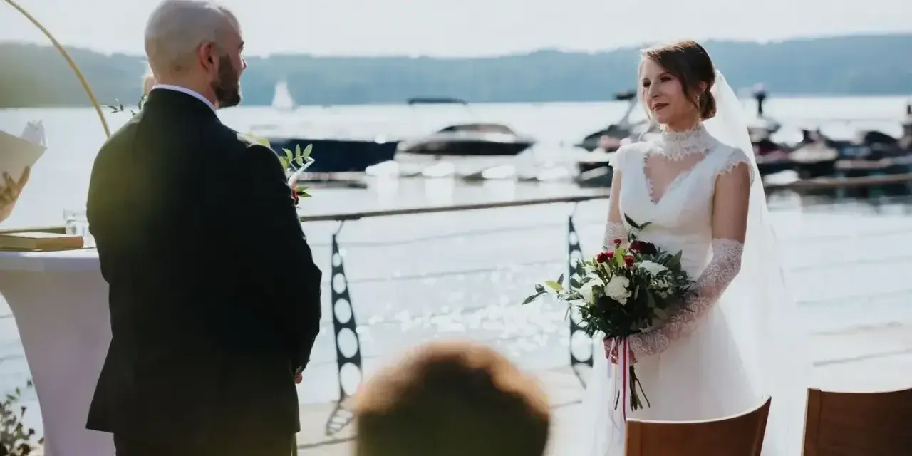 Para Młoda podczas ceremonii ślubu kanonicznego nad jeziorem. Ona w białej sukni z bukietem, on w garniturze.
