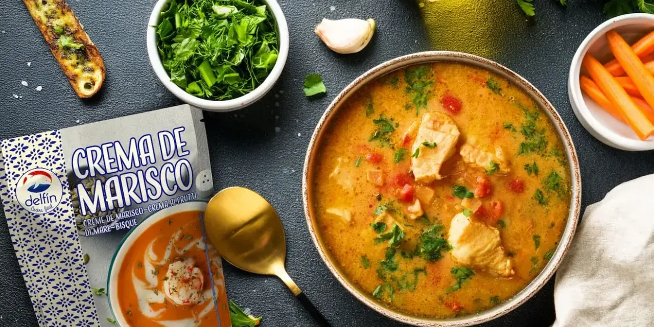 Un tazón de sopa de pescado casera con trozos de pescado, tomates y perejil, junto a una tostada, zanahorias y un paquete de crema de marisco.
