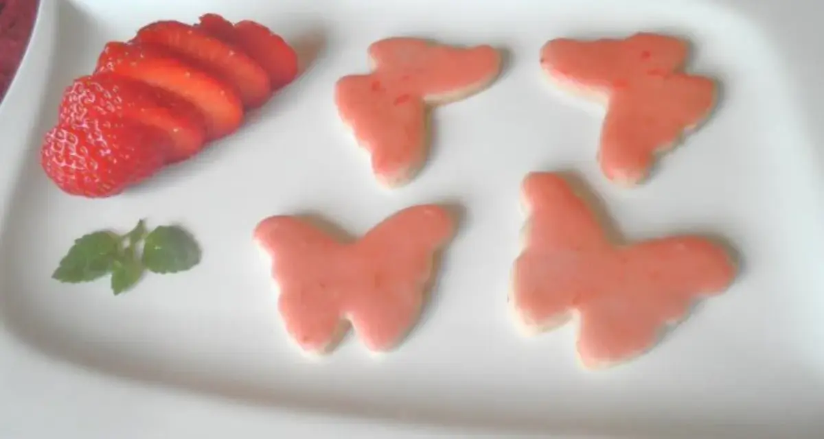 Zarte Erdbeer-Kekse in Schmetterlingsform, dekoriert mit rosa Zuckerguss, neben frischen Erdbeerscheiben und einem Minzblatt.