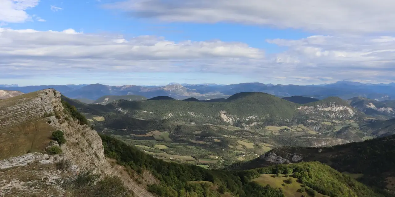 Vue panoramique depuis le col de Perty, avec des montagnes verdoyantes et un ciel bleu parsemé de nuages.