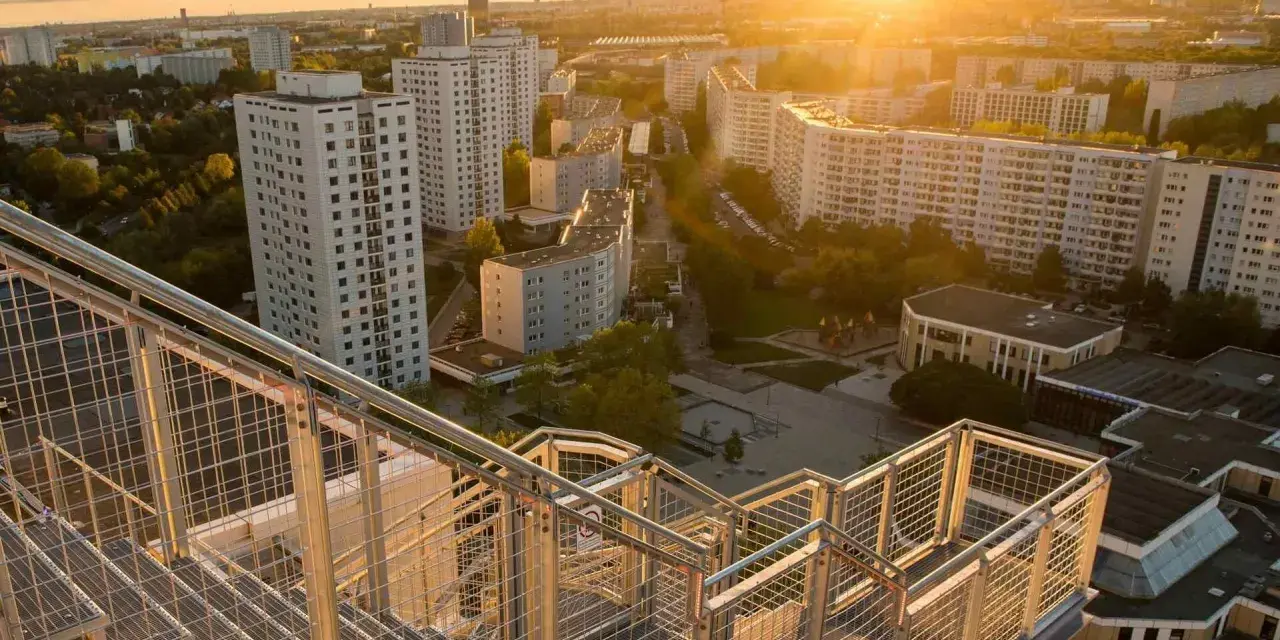 Blick von einer Treppe auf den Skywalk Marzahn bei Sonnenuntergang. Hohe Wohnhäuser und grüne Flächen prägen die Szenerie.